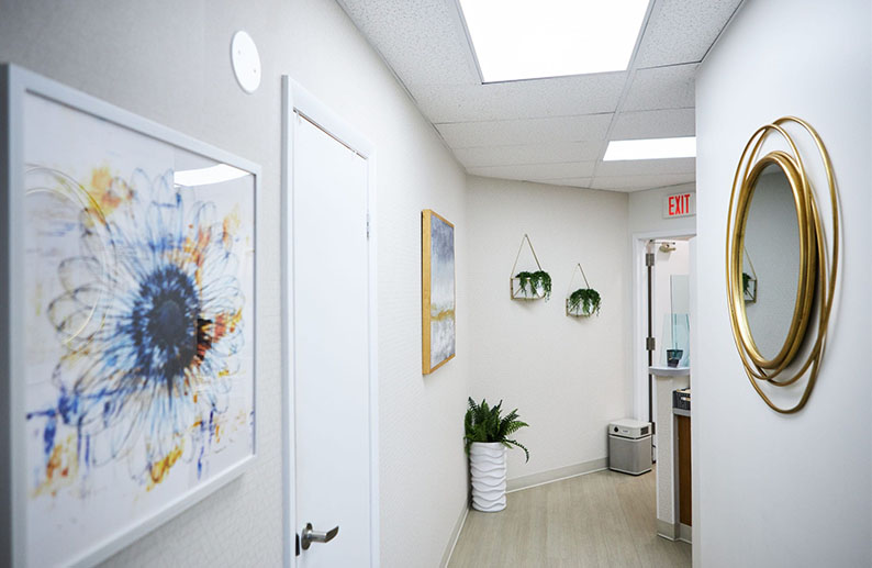 The image shows an interior space, likely a waiting area or hallway within a medical office, featuring artwork on the walls, a circular mirror, and a door with a round window.